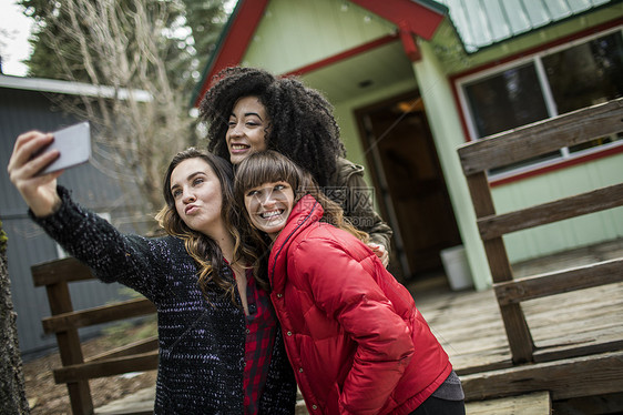 三个朋友站在小木屋旁边拿着自拍使用智能手机图片