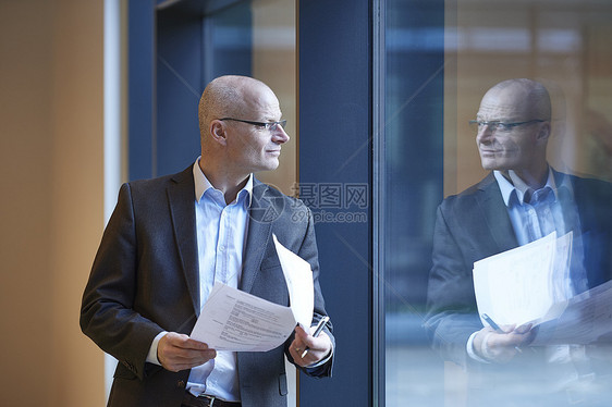 拿着文件通过办公室窗口看向窗外的成熟商人图片