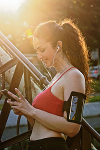 年轻的女跑青年戴耳机在阳光下看智能手机图片