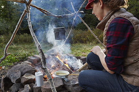 坐在美国纽约州卡茨基尔公园高露洁湖野生森林营火旁烹饪食物的妇女图片