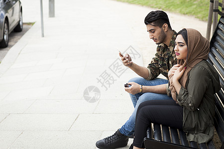 坐在公园长椅上的年轻夫妇男人看着手机图片