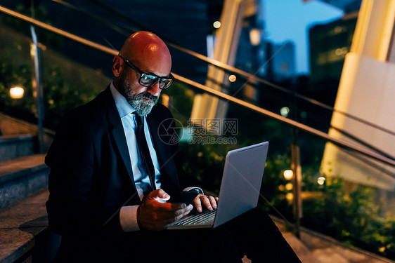 成熟商人夜间户外坐在台阶上使用笔记本电脑持有智能手机图片