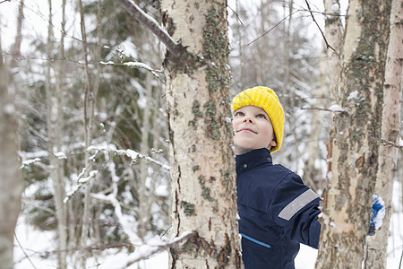 戴着黄色针织帽的男孩仰望着被雪覆盖的树图片