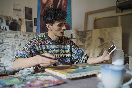男艺术家在艺术家工作室画画布时看着智能手机图片