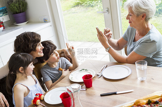 祖母坐在厨房用智能手机拍摄成年女儿和孙子的照片图片