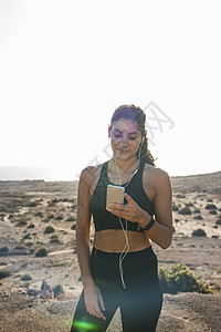 在西班牙加那利群岛拉斯帕尔马的干旱沿海景观中寻找智能手机的年轻女跑者图片