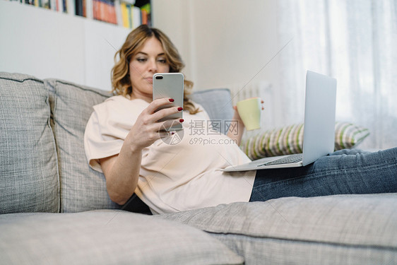 在沙发上腿上放着笔记本电脑的孕妇在玩手机图片