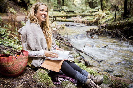 年轻女子拿着笔记本在森林河岸上微笑背景图片