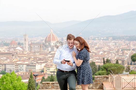 以意大利托斯卡纳佛罗伦萨市为背景的年轻夫妇看着手机上微笑图片