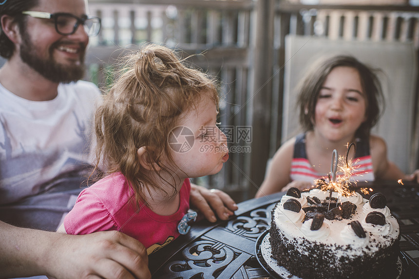 与姐妹和父亲一起的女幼儿在庭院桌上的庆祝蛋糕吹出火花图片