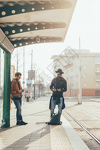 两个年轻的时尚男性看着智能手机在城市电车站等候图片
