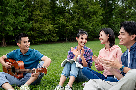 吉他弹奏不看镜头愉悦满意快乐的大学生在草地上弹吉他唱歌背景