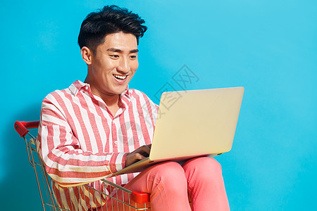 时尚网络购物微笑青年男人坐在购物车里用笔记本电脑图片