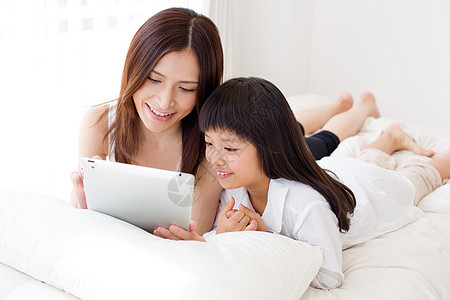 母亲陪着小女孩在床上看平板电脑图片