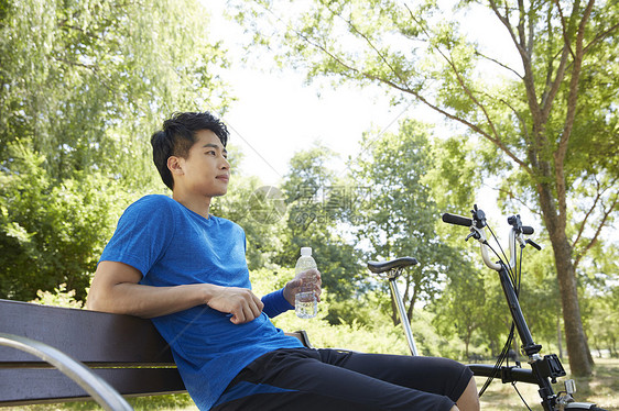坐在公园长凳上休息喝水的男青年图片