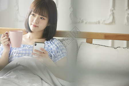 床上拿着水杯和手机的年轻女子图片