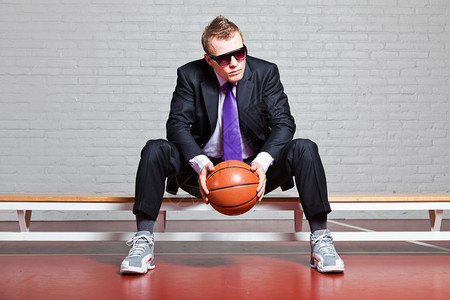 与篮球的商人戴着墨镜很帅与短的金发的年轻人坐在长凳上在室内体育馆图片