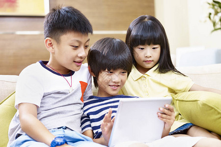 三名亚裔儿童坐在沙发上在家里使用数字平图片