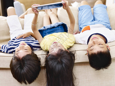 三名亚洲儿童躺在沙发上在家里使用数字平图片