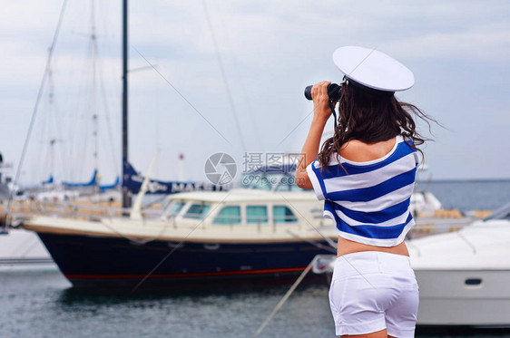 女孩通过双筒望远镜在海港看身着海洋风格的女人站图片