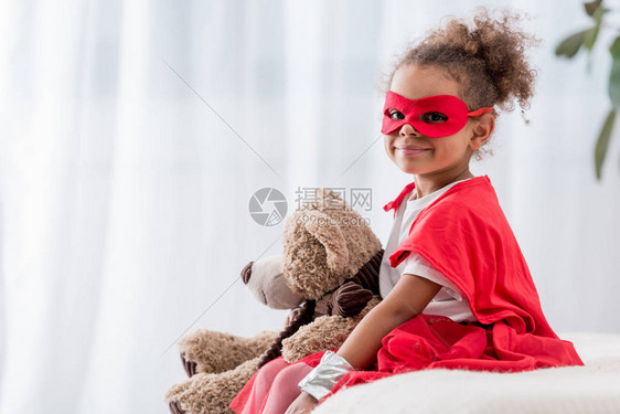 身着超级英雄服装和面具的可爱的非洲小孩带着泰迪熊图片