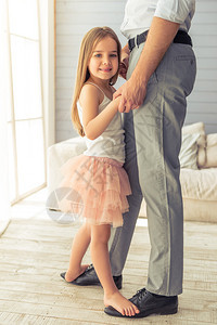 裁剪的年轻父亲和他可爱的小女儿在家里跳舞的形象女孩站在她父亲的脚上图片