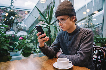 年轻英俊的时髦男人在靠近窗户的一家不错的咖啡馆里使用手机茶在桌子上图片