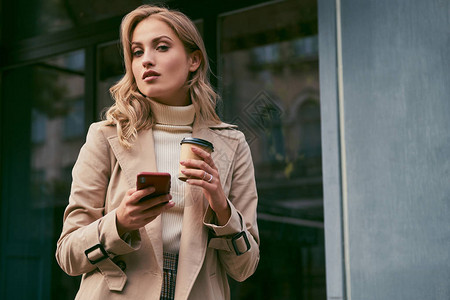 穿着海沟大衣的美丽散心金发美女喝着咖啡到外面去手机自信地在户背景图片