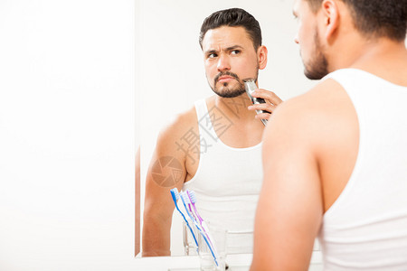 英俊的年轻人用电动剃刀在浴室镜子前剪胡须图片