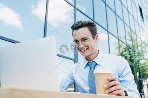 利用笔记本电脑和在现代建筑外持有纸杯的眼镜上装着笑得英俊笑着微图片