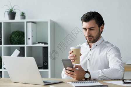 在工作场所使用智能手机用纸杯咖啡机的英俊青年商人手图片