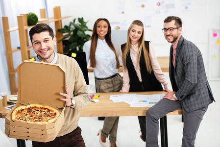 手拿披萨的微笑商人和办公室背后的多民族同事图片