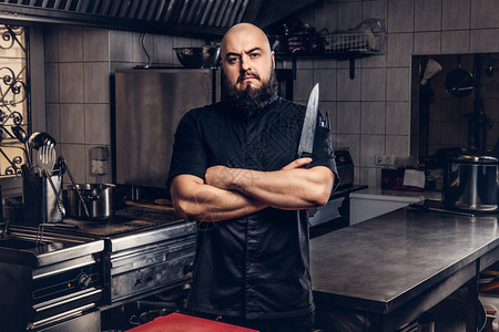 穿黑制服的胡子厨师拿着刀站在厨房里背景图片