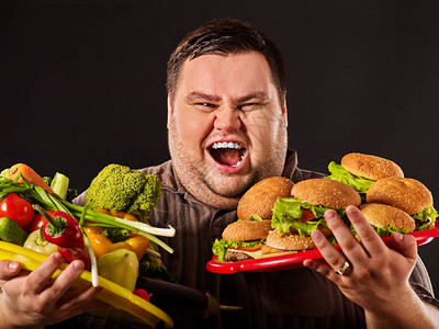 在健康和不健康的食物之间做出选择的节食胖子第一次在黑色背景下尝试减肥的超重男与汉堡包和蔬菜托盘为图片
