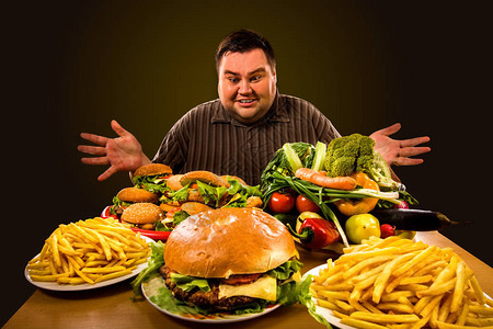 在健康和不健康的食物之间做出选择的节食胖子超重男第一次尝试减肥时吃汉堡包炸薯条和蔬菜托盘图片