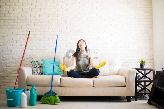 年轻家庭主妇穿戴黄色橡皮手套和清洁设备坐在沙发上静坐以缓解压力图片