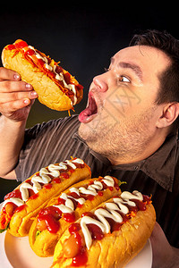胖子在盘子上吃快餐热狗超重者的早餐垃圾餐会导致肥胖人经常在黑色背景上暴饮食背景图片
