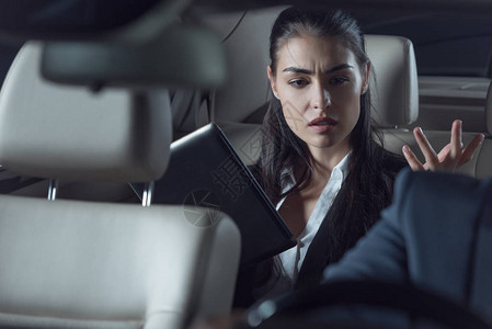 穿着西装的年轻不高兴女人坐在汽车后座与数字平板电脑和责骂司机图片