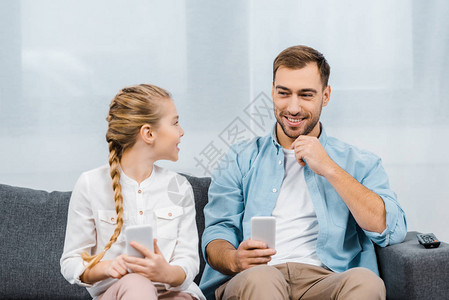 微笑的父女坐在沙发上拿着智能手机在客图片