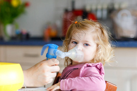 蹒跚学步的小女孩在家里用雾化器吸入帮助和拿着设备的父亲或母亲孩子有流感咳嗽和支气管炎哮喘吸入器吸图片