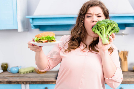 超重女人的肖像汉堡肉咬新鲜西兰花在家中厨房里手握着图片
