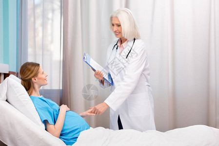 以剪贴板和听诊器与躺在医院床上的孕妇交谈的方图片