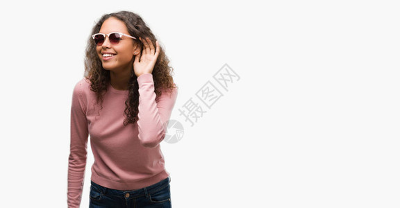 美丽的年轻西班牙裔妇女戴着太阳镜微笑着用手捂着耳朵听谣言或八卦图片