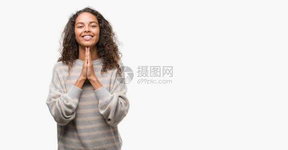 美丽的年轻西班牙女人穿着条纹毛衣用双手一起祈祷祈求宽恕微笑着满怀信图片