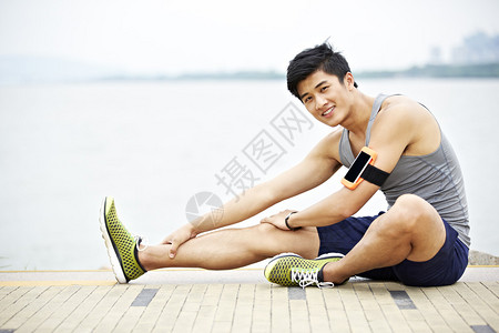 身着健身追踪器的年轻英俊年轻的年青亚洲慢跑运动员在湖边伸展一条腿图片