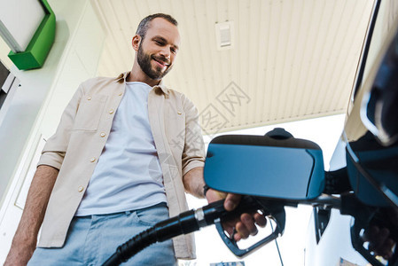 在加油站为汽车加油时持有燃料泵的快乐男子低角度视图图片