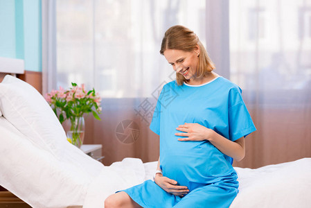 快乐的天主教孕妇摸着肚子坐在医院的床图片