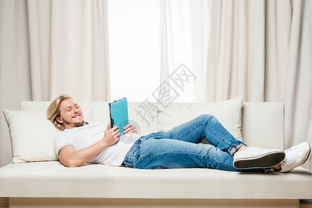 穿着白色T恤衫和牛仔裤的男子在家中躺在沙发上时使用数字图片