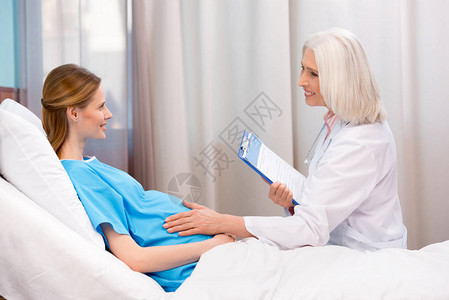 持有剪贴板和与躺在医院床上的孕妇交谈的高级医生图片