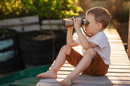 通过双筒望远镜在河岸上看的小男孩的画像带双筒望远镜的赤脚孩子坐在图片